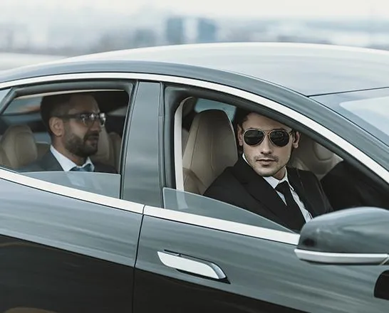 A driver in sunglasses at the wheel of a car, with a passenger behind